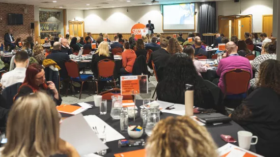 A large room with people at round tables listening to a Centrepoint speaker on the stage