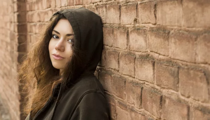 Young person with long brown curly hair in a brown hoodie standing against a brick wall