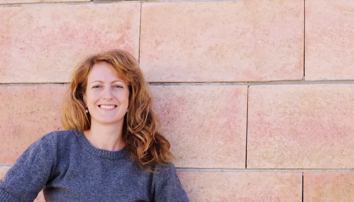 Person with long red hair wearing a blue jumper stands in front of a pink tiled wall.