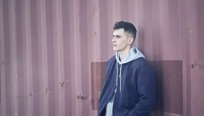 Young person in blue jacket and grey hoodie stands against a corrugated metal wall