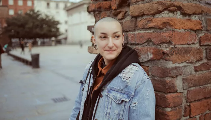 Avery stands against a brick wall, looking over their shoulder towards the camera with a half smile. They wear a denim jacket, brown hoodie and orange shirt.