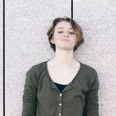 Young person with short hair leans against a grey wall 