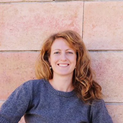 Person with long red hair wearing a blue jumper stands in front of a pink tiled wall.