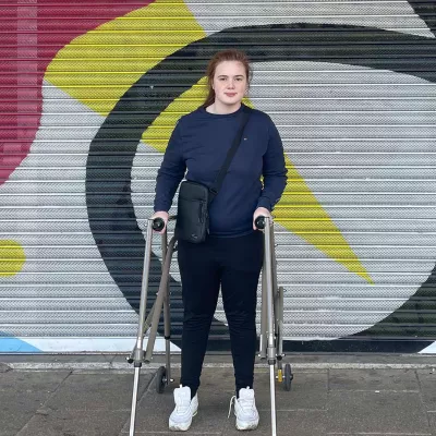 Young person with a walking frame in front of a colourful shop shutters