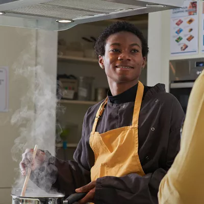 A young person stands in an apron while cooking food in a saucepan on a stove